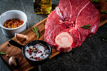 Raw meat with ingredients for cooking. Beef fillet, tenderloin on bone, on a cutting board, with salt, pepper, parsley, rosemary, olive oil, garlic, spices. On a black stone table, copy space