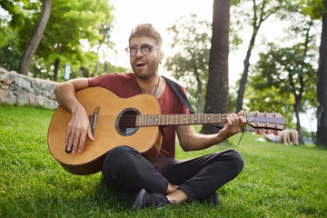 Stylish guy with pierced ear wearing trendy sunglasses and red t-shirt singing songs and playing guitar, sitting on grass in park.