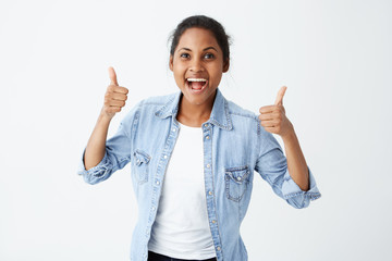 Glad young dark skinned female wearing denim long sleeved shirt making thumbs up sign and smiling cheerfully, showing her support and respect to someone. Body language. Good job.