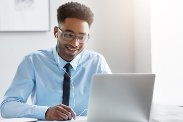 Happy businessman wearing elegant eyewear and shirt watching video online while sitting in front of opened laptop in earphones. Dark-skinned male communicating online with his friends, smiling gently