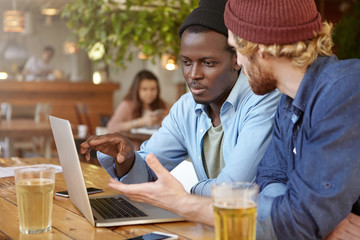 Two friends being at coffee shop, having drinks and discussing something in front of opened laptop, trying to make important decision. Mixed race hipster men meeting together for making project