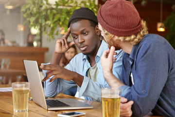 Two mixed race men colleagues chatting together indoors with cold drinks, looking attentively in screen of laptop, discussing business plans. Black male and his friend working together with computer