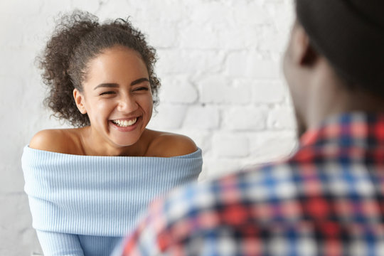 Cheerful mixed race female with curly hair sitting opposite her best African friend, having pleasant conversation with him, being glad to see him. Two black friends having fun together at cafe - Powered by Adobe