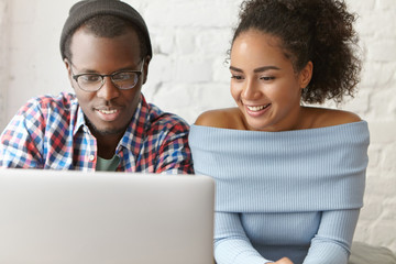 Two dark-skinned male and female sitting next to each other, looking attentively in screen of laptop, watching films or funny video together using free internet connection. Friendship concept