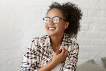 Joyful mixed race woman with curly pony tail wearing casual checkered shirt smiling pleasantly and closing her eyes with excitement, feeling relaxation and joy while being glad to meet with friends