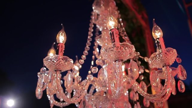 Candle-like, glass crystal, chandelier ceiling lights on the metal frame. On the backgroung - light of the latern and night sky. Romantic evening. Wedding decor. Twilight. Steadycam. Slowly going away