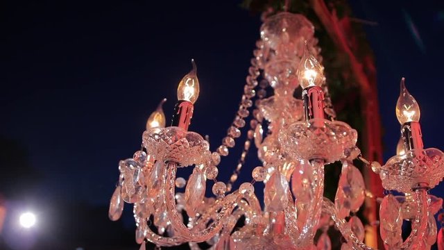 Candle-like, glass crystal, chandelier ceiling lights on the metal frame. On the backgroung - light of the latern and night sky. Romantic evening. Wedding decor. Steadycam. Slowly closing up and