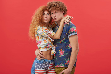 Cropped shot of charming young woman with freckles and long wavy hair dressed in summer American flag print denim shorts and floral design top holding hands tight around her handsome boyfriend