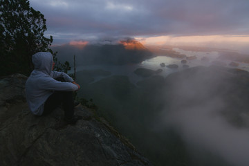 Man sitting on the edge with campfire above bay with islands and forests in cloudy weather from top view in the evening