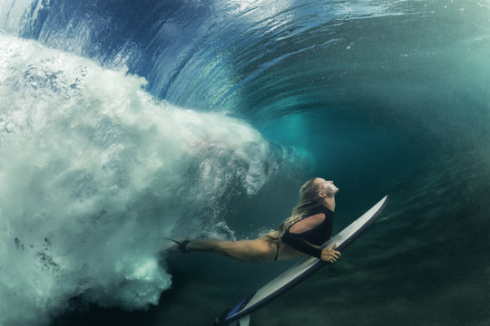 A blonde surfer girl underwater doing duck dive holding surfing board left behind air bubbles in blue water background under big ocean wave