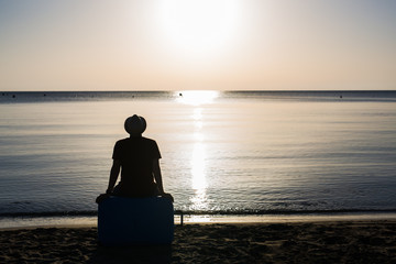 Back view silhouette of man sitting on suitcase on blue sea outdoors background. Arriving at...