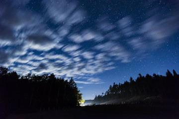 Night sky full of stars and swift moving clouds above a wilderness landscape and road, car headlights