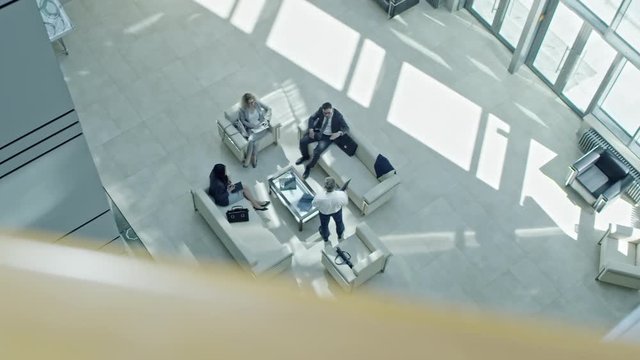 PAN with top view of gesturing businessman presenting magazine to male and female coworkers sitting on sofas and armchair in lobby of multi-storey business centre with panoramic windows