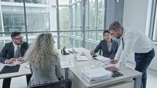 PAN of businessman in shirt and tie showing document to Asian businesswoman in glasses during meeting with colleagues in office with glass walls