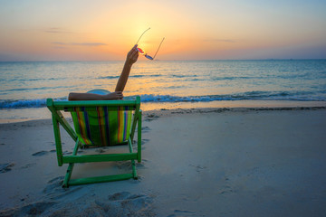 woman lie down on the deck chair at rim of the sea beach, hand up with sunglass at sunset into the...