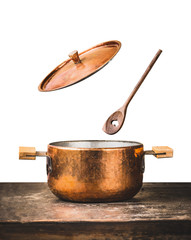 Copper cooking pot with flying  open lid and wooden spoon on table, isolated on white background,...