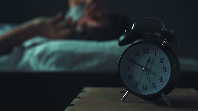 Adult caucasian man talking on mobile phone late at night while lying in bed in bedroom, selective focus on alarm clock on bedside table