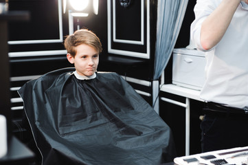 Young handsome barber making haircut of cute boy in barbershop