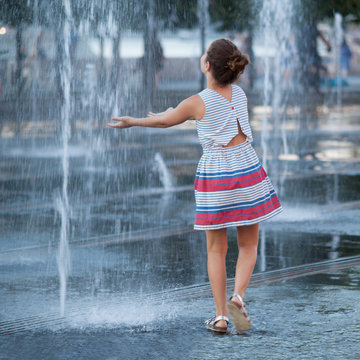 A teenage girl walks by the jets of a fountain and stretches out her arms trying to catch a spray