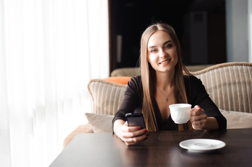 beautiful girl drinking tea and using a mobile phone while sitting in a cafe in front of a large window