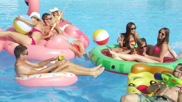 SLOW MOTION CLOSE UP Happy smiling students have water gun fight on colorful floaties at pool party on spring break. Cheerful young people have fun on inflatable flamingo pizza watermelon and doughnut