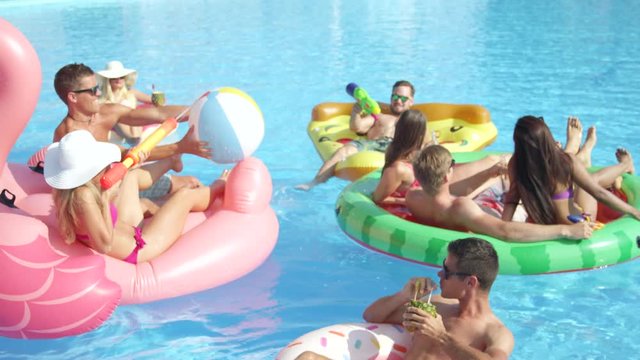 SLOW MOTION CLOSE UP: Playful smiling friends enjoying hot summertime on colorful floaties at pool party. Cheerful young people having fun on inflatable pizza, flamingo, watermelon and doughnut floats