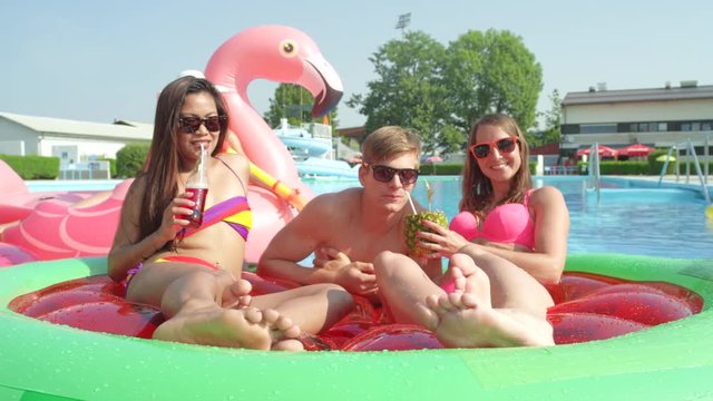 SLOW MOTION CLOSE UP: Smiling young girls and guys drinking alcoholic cocktail drinks on fun inflatable watermelon and flamingo floats. Cheerful friends enjoying summer on colorful floats in pool
