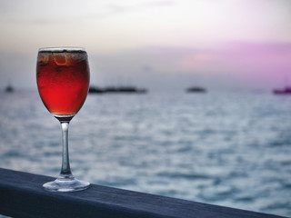 Red iced cold beverage in wine glass on wood piece with sunset background of open sea and silhouette boats under pale orange and pink sky. This image was taken at a tropical resort in Maldives island