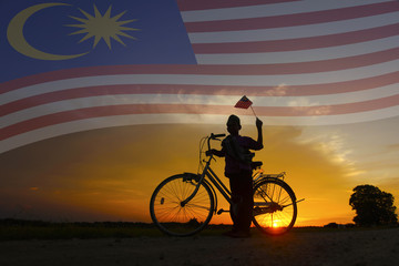 Naklejka premium Independence Day concept - Silhouette of young local boy on paddy field holding a Malaysian flag during sunset.