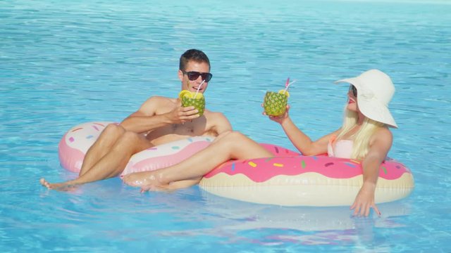 SLOW MOTION: Young couple drinking pineapple cocktails and relaxing on fun pink doughnut floaties in pool. Young man and woman in love relaxing on inflatable doughnut floats on sunny summer vacation