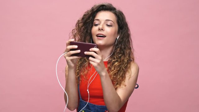 Young happy caucasian woman play games by phone isolated over pink background