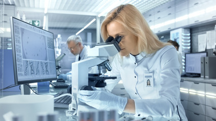 Female Research Scientist Looks at Biological Samples Under Microscope. She and Her Colleagues Work in a Big Modern Laboratory/ Medical Centre. - obrazy, fototapety, plakaty
