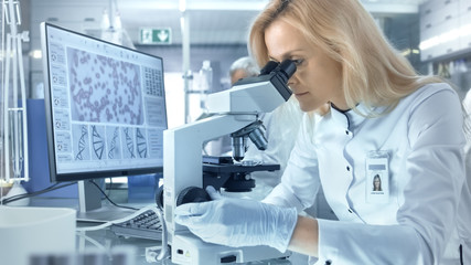 Female Research Scientist Looks at Biological Samples Under Microscope. She and Her Colleagues Work in a Big Modern Laboratory/ Medical Centre.