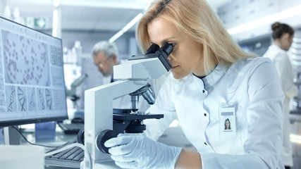 Female Research Scientist Looks at Biological Samples Under Microscope. She and Her Colleagues Work in a Big Modern Laboratory/ Medical Centre.