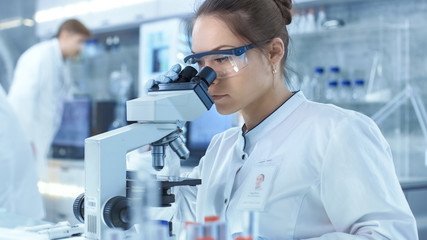 Medical Research Scientists Looking at Samples Under Microscope. She Works in a Bright Modern...