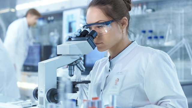 Medical Research Scientists Looking at Samples Under Microscope. She Works in a Bright Modern Laboratory.