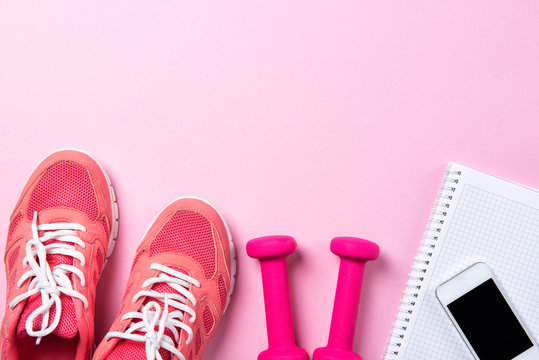 Fitness concept, pink sneakers and dumbbells with notebook with smart phone on pink background, top view with copy space