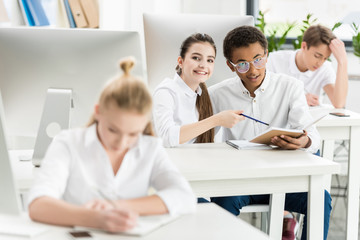 selective focus of multicultural smiling students looking at camera during lesson in classroom