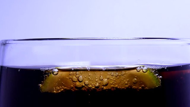 Pouring Soda and Slices of Lime. Brown Soda Pop In Glass. Releasing Bubbles And Foaming In Parkling Water. Aerated Powder Water Is Drawn into Glass On Background of White Wall In Isolated
