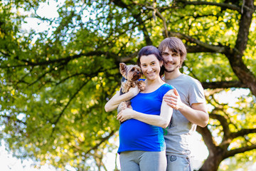 Fototapeta premium Man and pregnant woman in sports wear hugging and holding yorkshire terrier in hands after workout in summer time in green park.