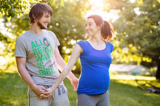 Bright picture of hipster man and pregnant woman in sports wear are holding by hands and looking at each other in beautiful summer day in green park.