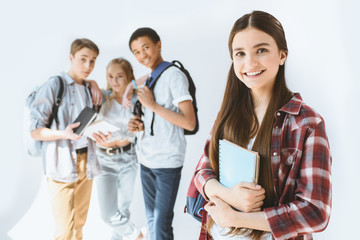 smiling teenage girl with notebook in hands looking at camera with multicultural students behind isolated on white