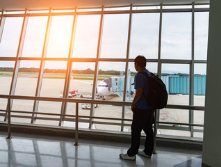 Man looking on the private jet,Asia young man is standing near window at the airport and watching plane before departure. He is standing and carrying luggage. Focus on his back,tourist,trip.