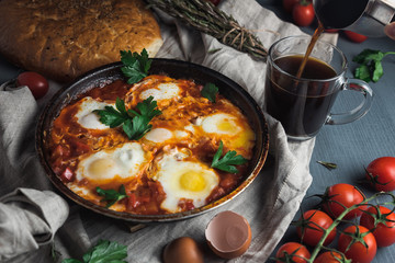 Shakshuka with pita bread in a pan. Fried eggs, onion, bell pepper, tomatoes and parsley on a rustic wooden table with ingredients.