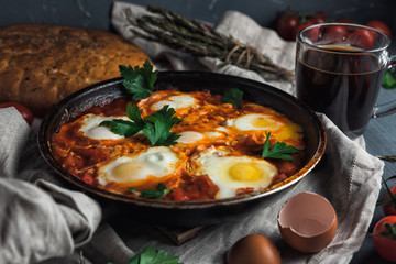 Shakshuka with pita bread in a pan. Fried eggs, onion, bell pepper, tomatoes and parsley on a rustic wooden table with ingredients.