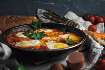 Shakshuka with pita bread in a pan. Fried eggs, onion, bell pepper, tomatoes and parsley on a rustic wooden table with ingredients.