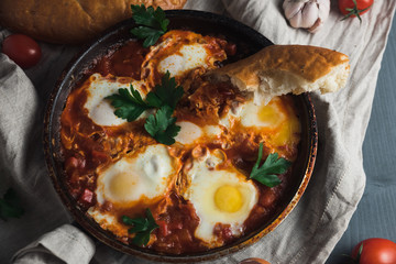 Shakshuka with pita bread in a pan. Fried eggs, onion, bell pepper, tomatoes and parsley on a rustic wooden table with ingredients.