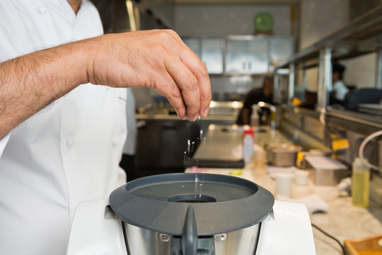 A hand dropping organic fresh rock salt into a food blending processor, in an industrial kitchen setting.