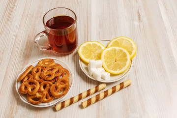 Transparent cup of black tea on a light table and saucers with pretzels, sweet sticks and chopped lemon, and pieces of refined sugar.