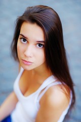 Fototapeta na wymiar Portrait of young serious girls long-haired brunette with beautiful brown eyes in a white t-shirt outdoors on gray-blue blurred background, closeup.